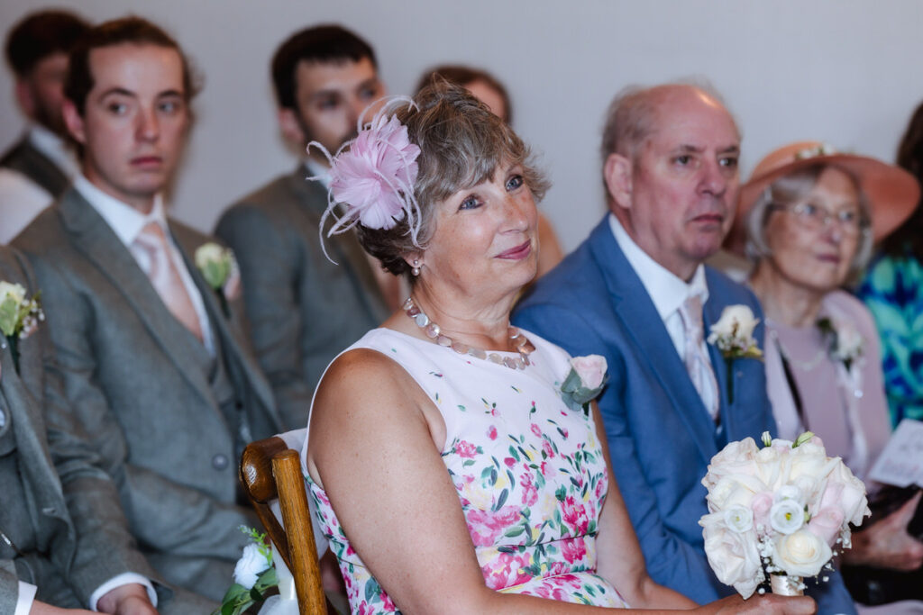 A woman in a floral dress and pink fascinator sits holding a bouquet, smiling at a wedding ceremony. Beside her are an older man in a blue suit and an elderly woman in a hat; other guests in suits are in the background.