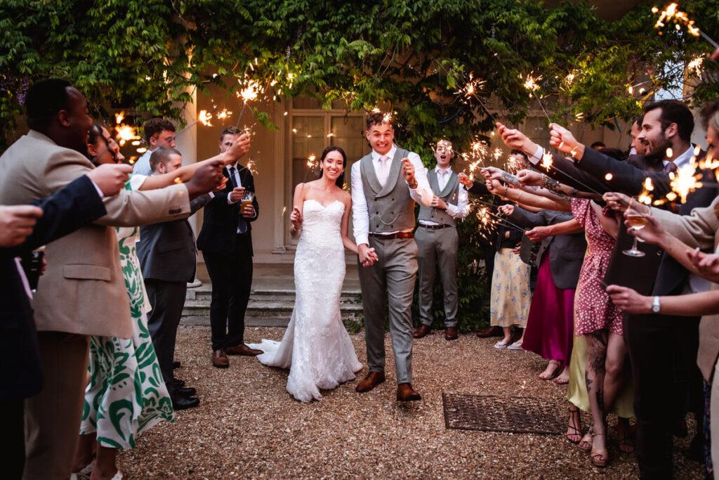 A bride and groom walk hand-in-hand outdoors, smiling, while guests on both sides hold sparklers and cheer. The scene is festive, with greenery and a building in the background.