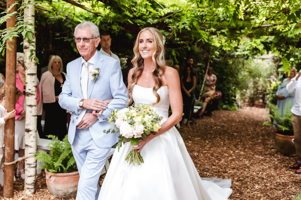 A bride in a white dress holding a bouquet walks down an outdoor aisle with an older man in a light blue suit. They smile, surrounded by greenery and guests seated on either side.