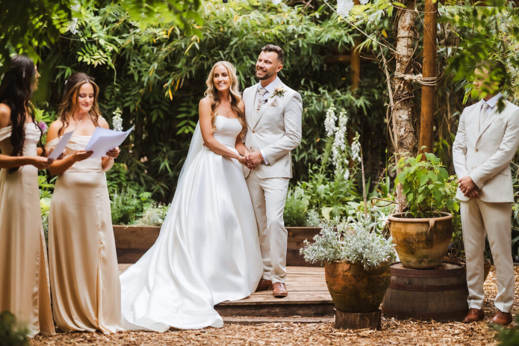 A bride and groom stand smiling and holding hands during an outdoor wedding ceremony, surrounded by greenery. Bridesmaids in light dresses and a groomsman in a light suit stand nearby.
