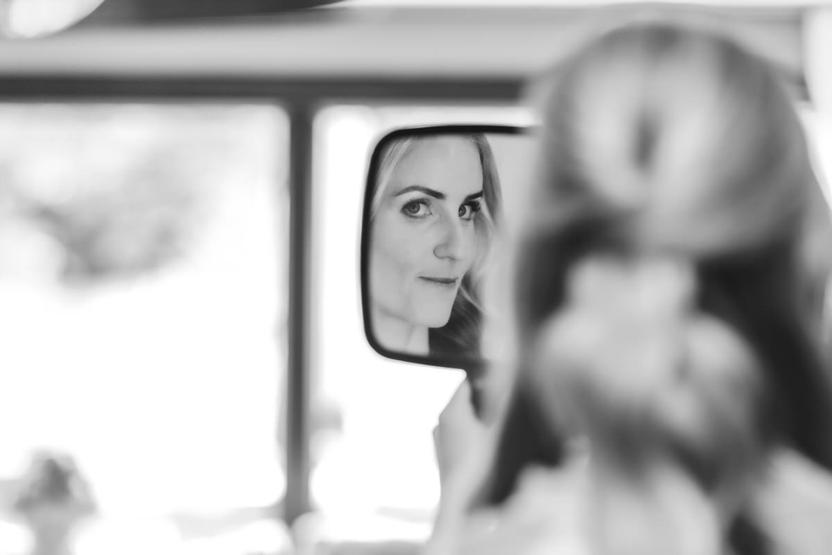 A woman with blonde hair is reflected in a side mirror, looking at the camera with a slight smile. The image is in black and white and the background is blurred.