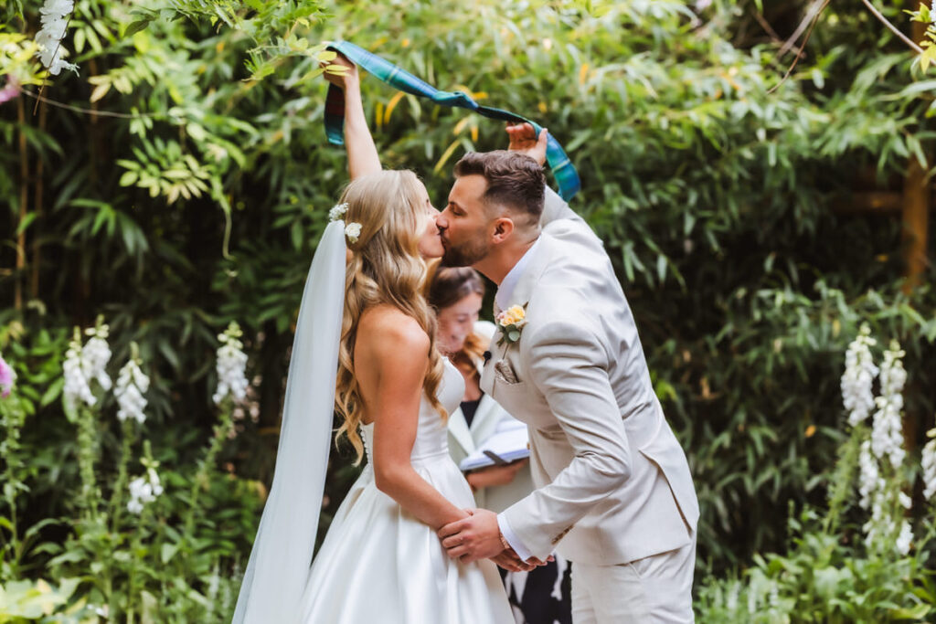 A bride and groom share a kiss during their outdoor wedding ceremony, holding hands as a celebrant drapes a ribbon over them, surrounded by lush greenery and flowers.