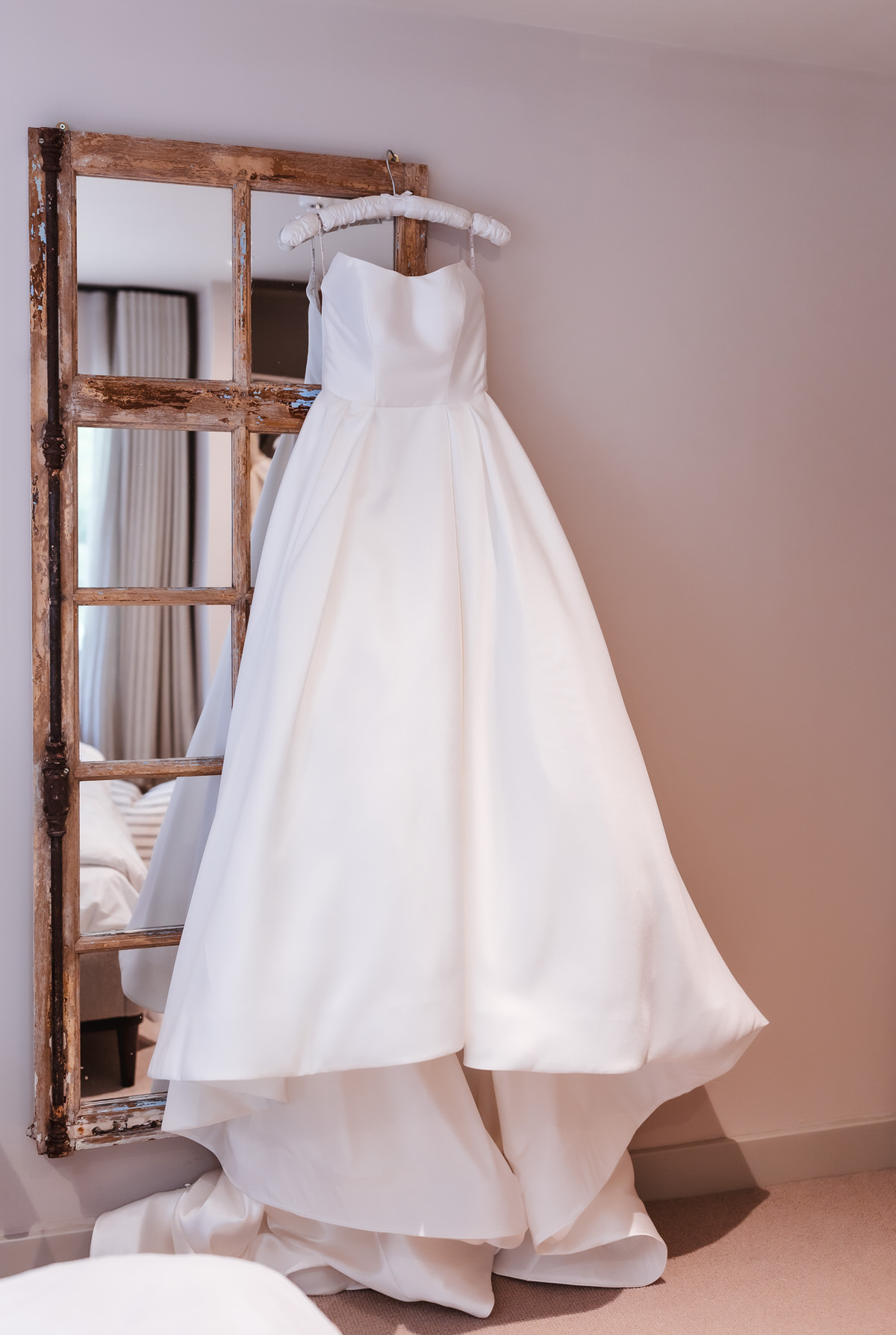 A strapless white wedding dress with a full skirt hangs on a rustic wooden-framed mirror in a softly lit room with beige walls and carpet.