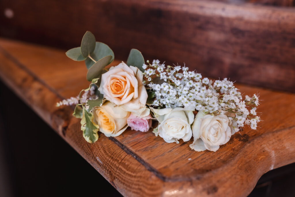 A small bouquet of roses in cream, yellow, and pale pink, accented with eucalyptus leaves and white babys breath, rests on a polished wooden surface.