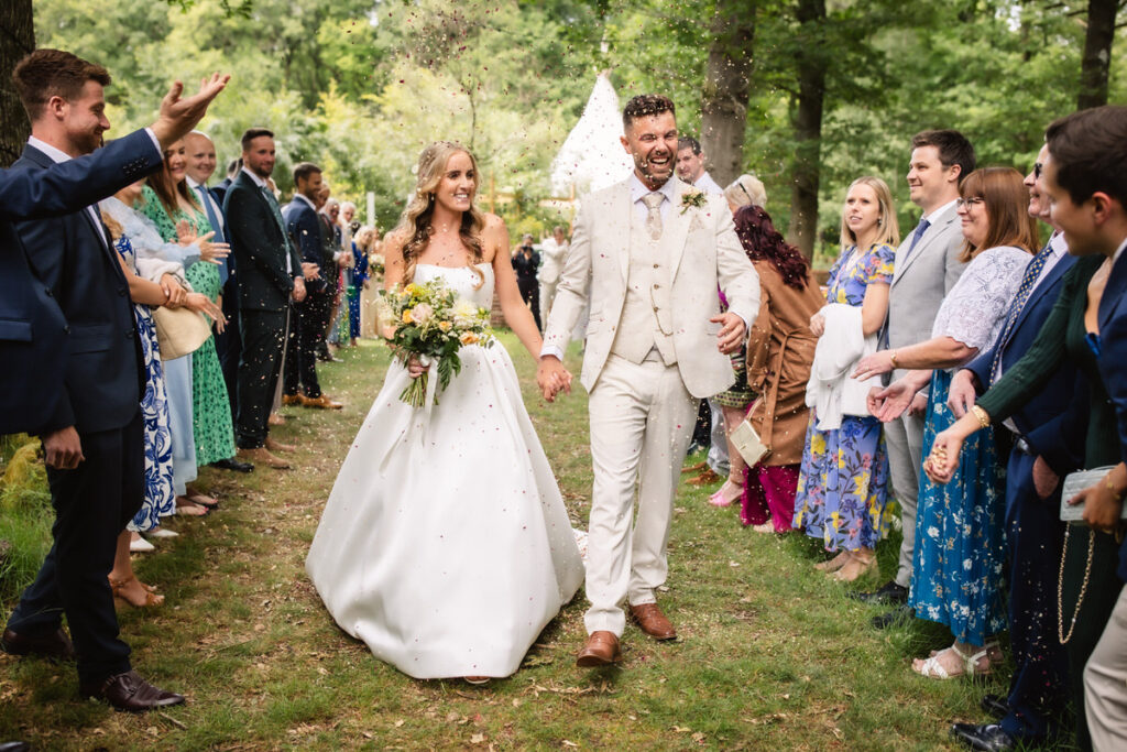 A bride and groom walk down an outdoor aisle, smiling, as guests on both sides throw confetti. The wedding takes place in a green, wooded setting, with people dressed in formal attire.