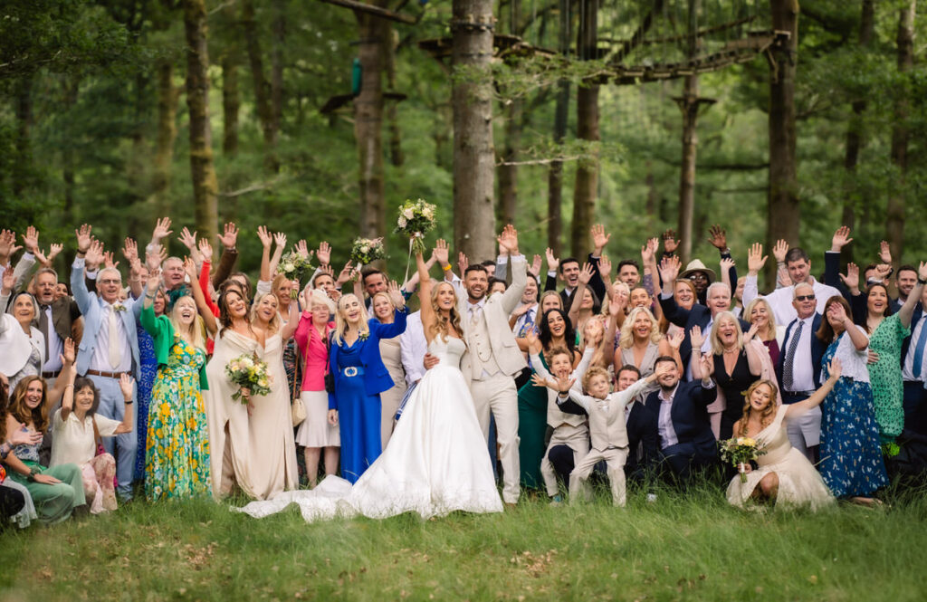 A large group of people, including a bride in a white gown and a groom in a light suit, pose and cheer with raised hands in a lush, green forest, celebrating a joyful outdoor wedding.