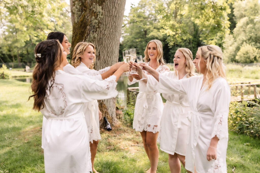 Six women in white robes stand outdoors under a tree, smiling and raising glasses in a toast. They appear to be celebrating, surrounded by greenery and sunlight.