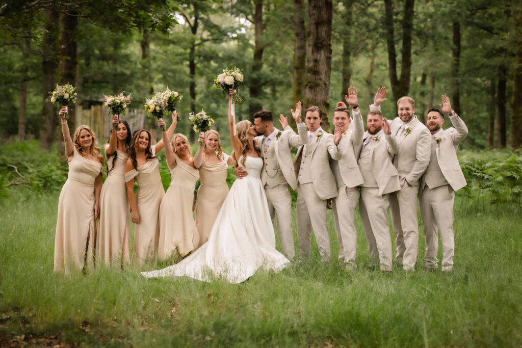 A bride and groom stand with their wedding party in a green forest. The bridesmaids wear light dresses and hold bouquets, while the groomsmen wear light suits. Everyone is smiling and some are raising their hands in celebration.
