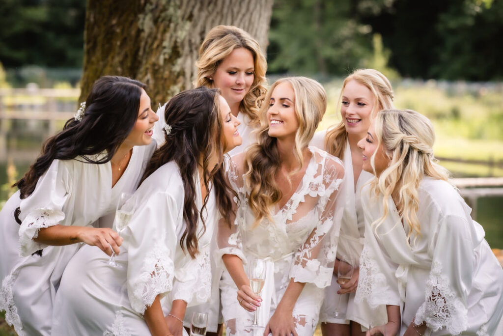 Six women in white robes, smiling and laughing together outdoors near a tree, holding glasses, with greenery and a fence in the background. The mood is joyful and celebratory.