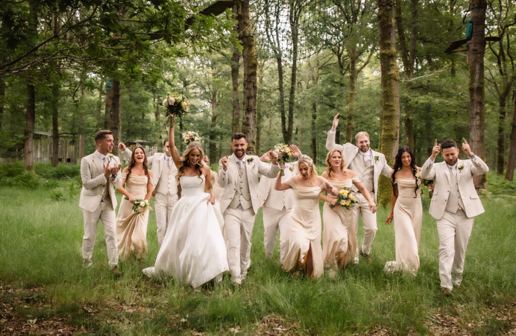 A joyful wedding party dressed in light suits and dresses walks through a green forest, laughing and raising bouquets in celebration. The group appears happy and energetic, surrounded by tall trees and lush grass.