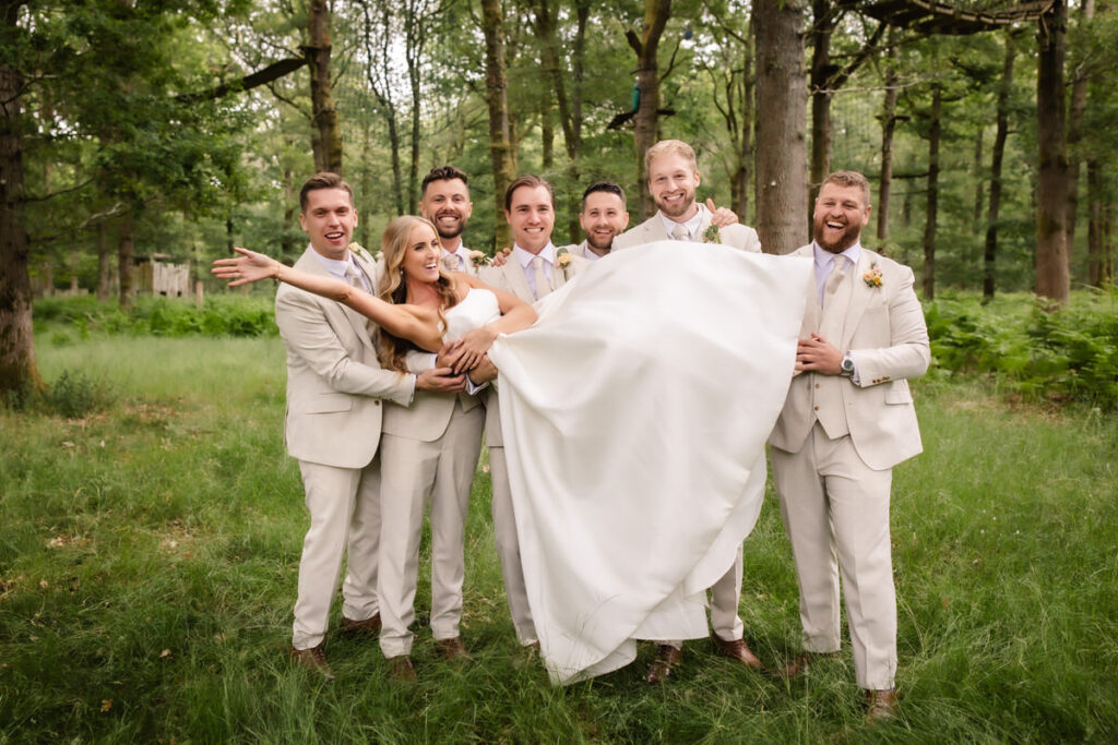 A bride in a white gown is held up horizontally by six groomsmen in light-colored suits, all smiling and laughing together in a green forest clearing.