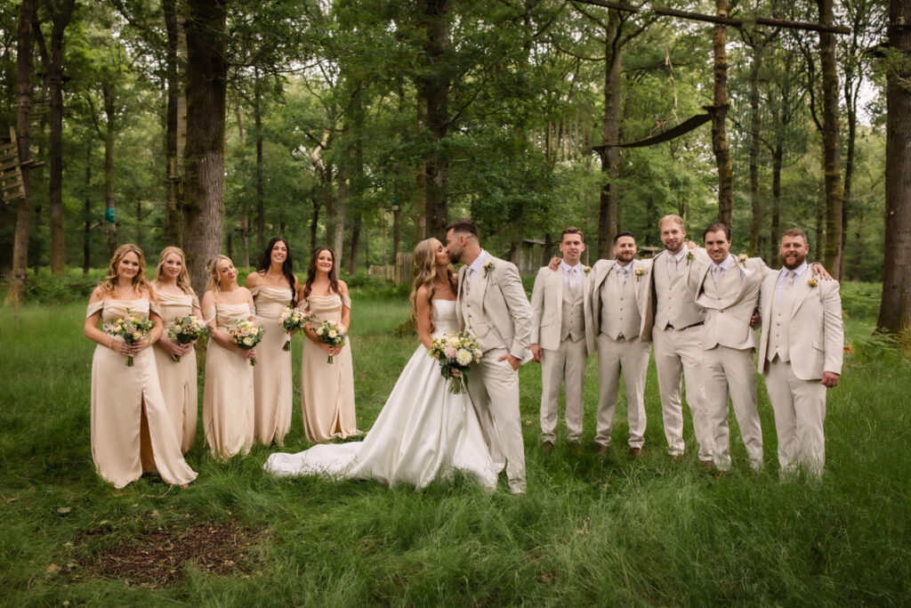 A bride and groom kiss in a forest clearing, surrounded by bridesmaids in light dresses and groomsmen in light suits, all smiling and holding bouquets.