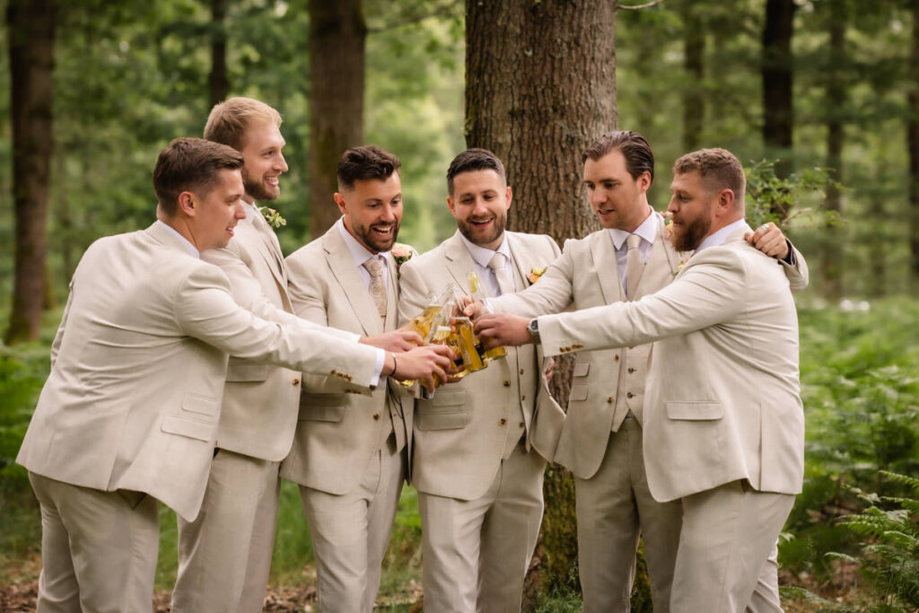 Six men in matching light beige suits stand in a forest, smiling and raising beer bottles together in a celebratory toast. Trees and greenery surround them, creating a relaxed, outdoor atmosphere.