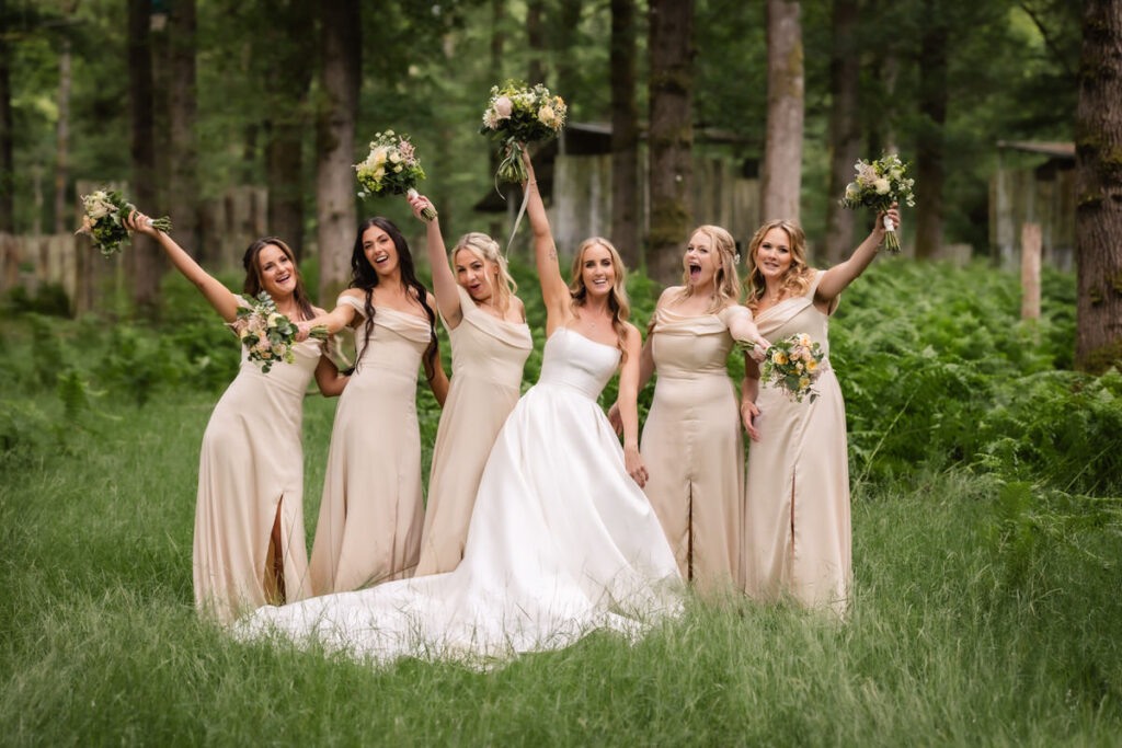 Six women pose joyfully in a lush, green forest. The bride wears a white gown, while the five bridesmaids in matching beige dresses hold bouquets and smile with raised arms.