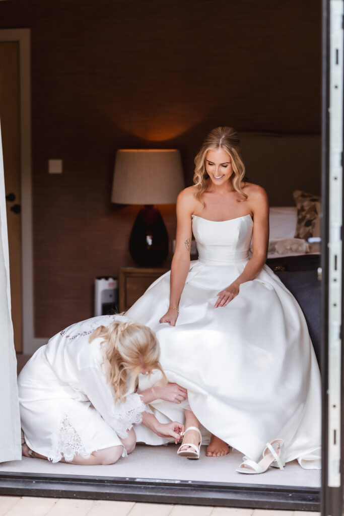 A bride in a white gown sits smiling on a bed while another woman in a robe kneels, fastening the brides white high-heeled shoe. The scene is indoors, with soft lighting and a lamp in the background.