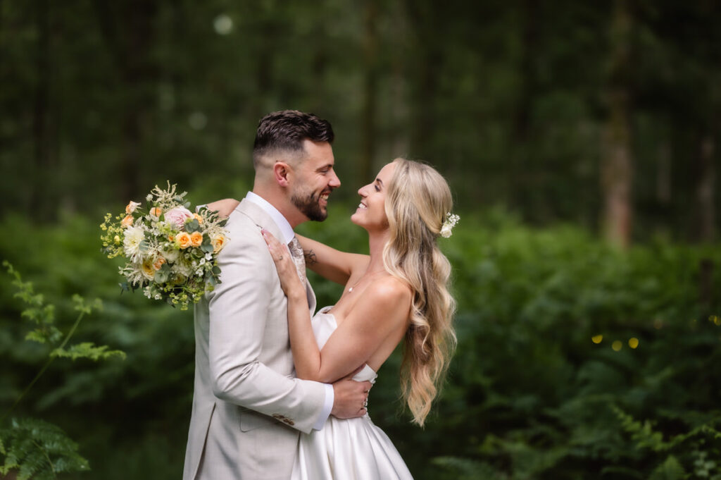 A newlywed couple embraces and smiles at each other in a lush green outdoor setting. The bride holds a bouquet of flowers and wears a white dress, while the groom wears a light-colored suit.
