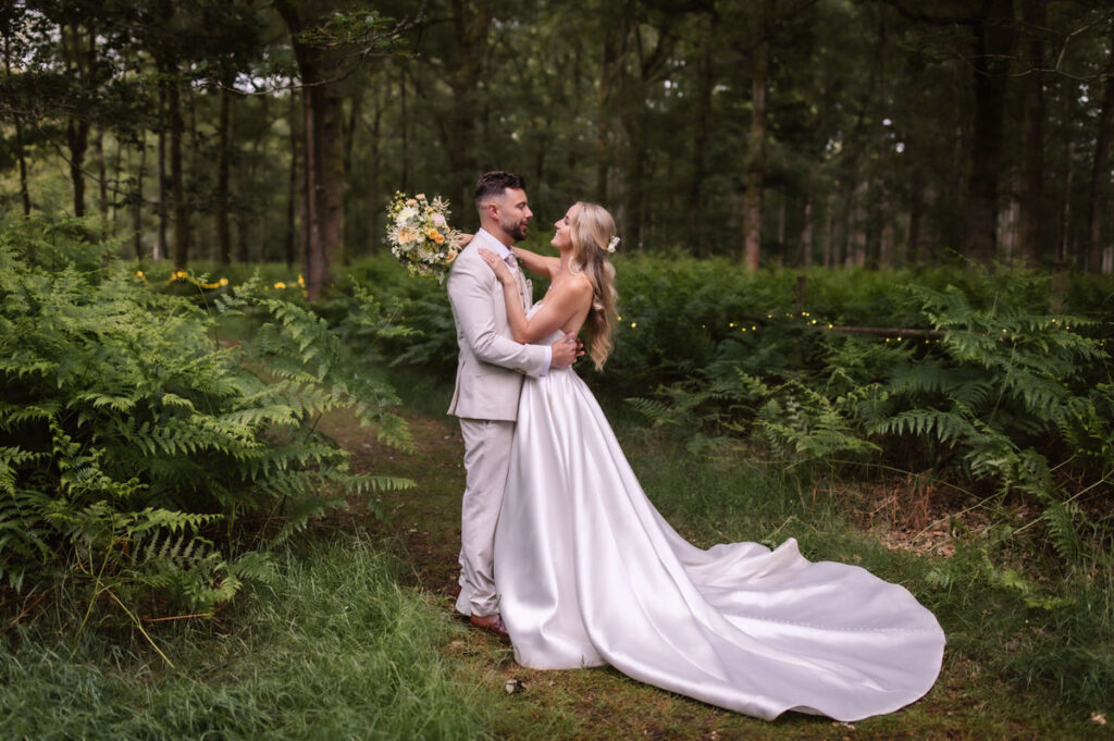 A bride in a long white gown and a groom in a light suit embrace and smile at each other in a lush, green forest setting, surrounded by ferns and trees.