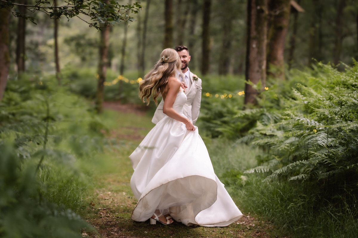 A bride and groom dance together on a forest path, surrounded by lush green ferns and trees. The bride’s white dress twirls as she spins, and the groom smiles, both dressed formally and appearing joyful.