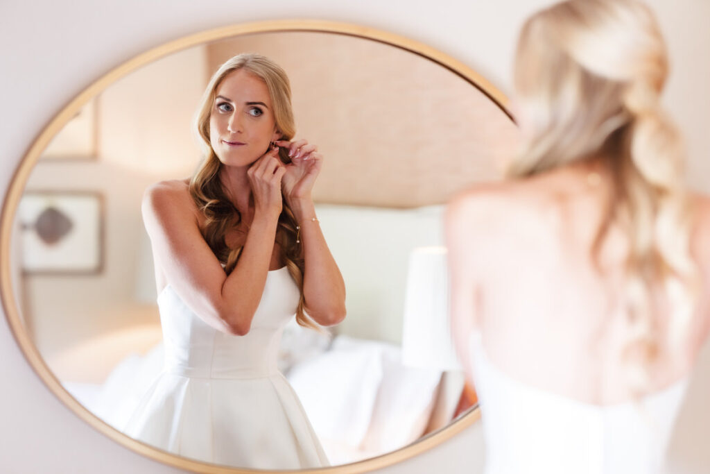 A woman in a white dress stands in front of a round mirror, adjusting an earring. Her long, wavy blonde hair falls over her shoulders, and the soft background suggests a calm, elegant setting.