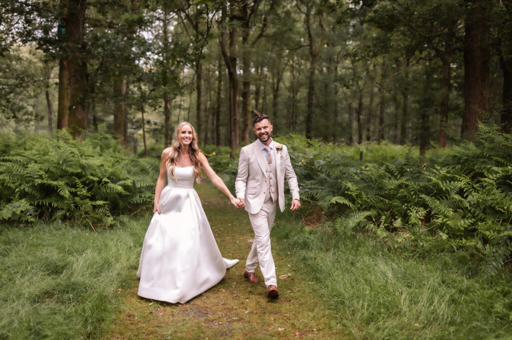 A bride in a white dress and a groom in a light suit walk hand in hand down a grassy path through a lush, green forest, both smiling joyfully.
