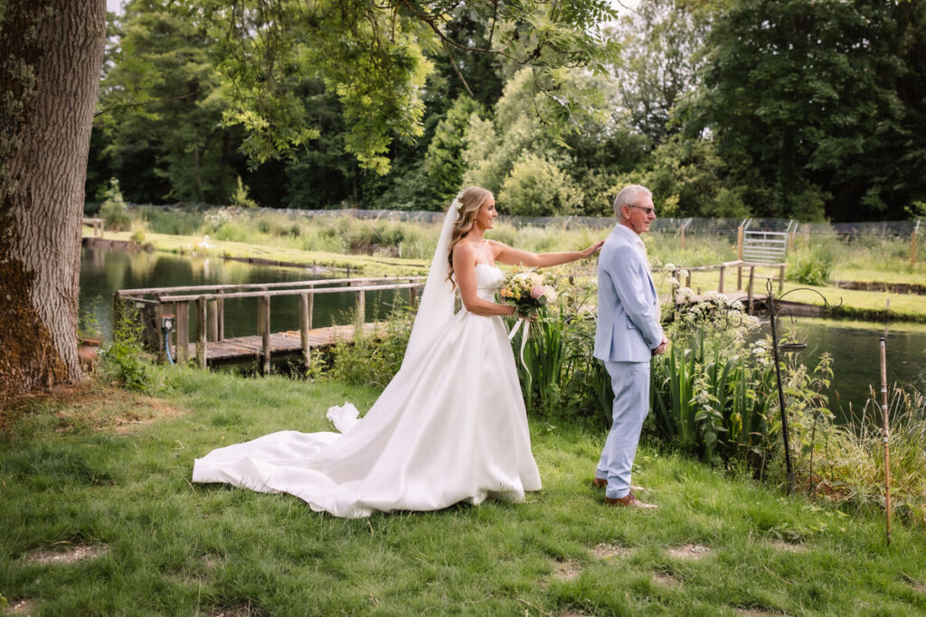 A bride in a white gown stands behind an older man in a light suit by a pond, about to tap him on the shoulder amid green grass, trees, and flowers.