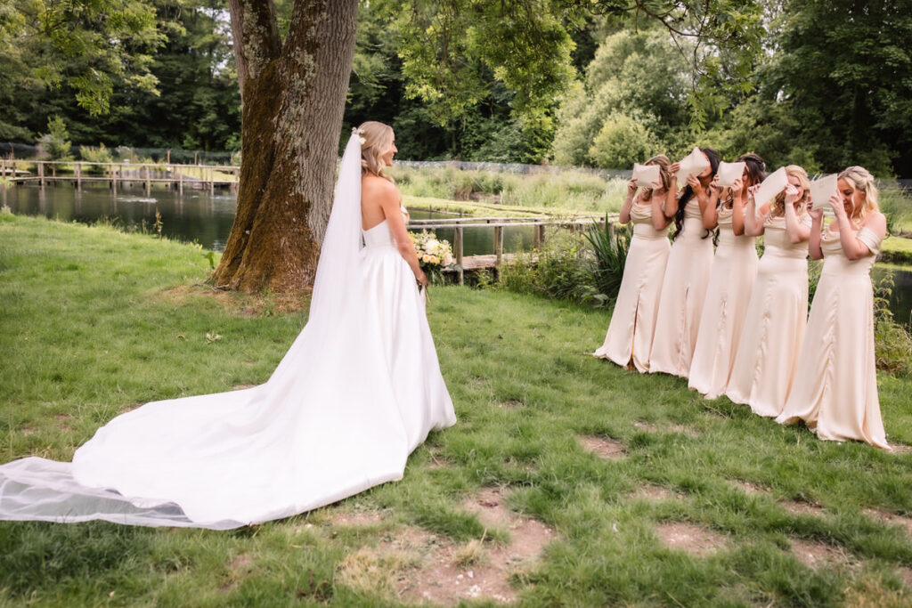 A bride in a white gown stands outdoors near a pond, facing six bridesmaids in matching cream dresses who are covering their faces with their hands in excitement or surprise.