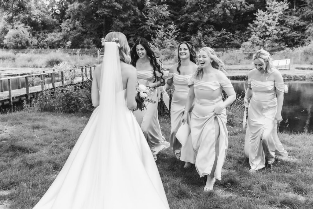 A bride in a long white gown and veil stands with her back to the camera as four bridesmaids in matching light dresses approach her, all smiling and laughing outdoors near a pond and greenery.