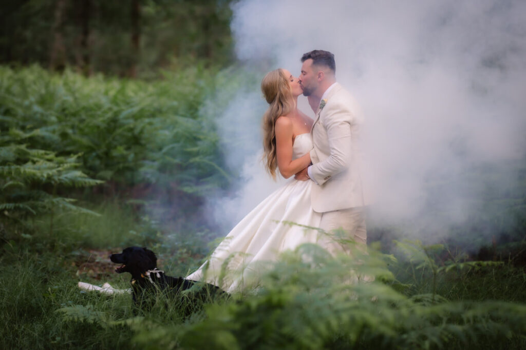 A bride and groom kiss in a lush green forest surrounded by mist, with a black dog lying in the grass nearby. The bride wears a white dress and the groom a light suit.