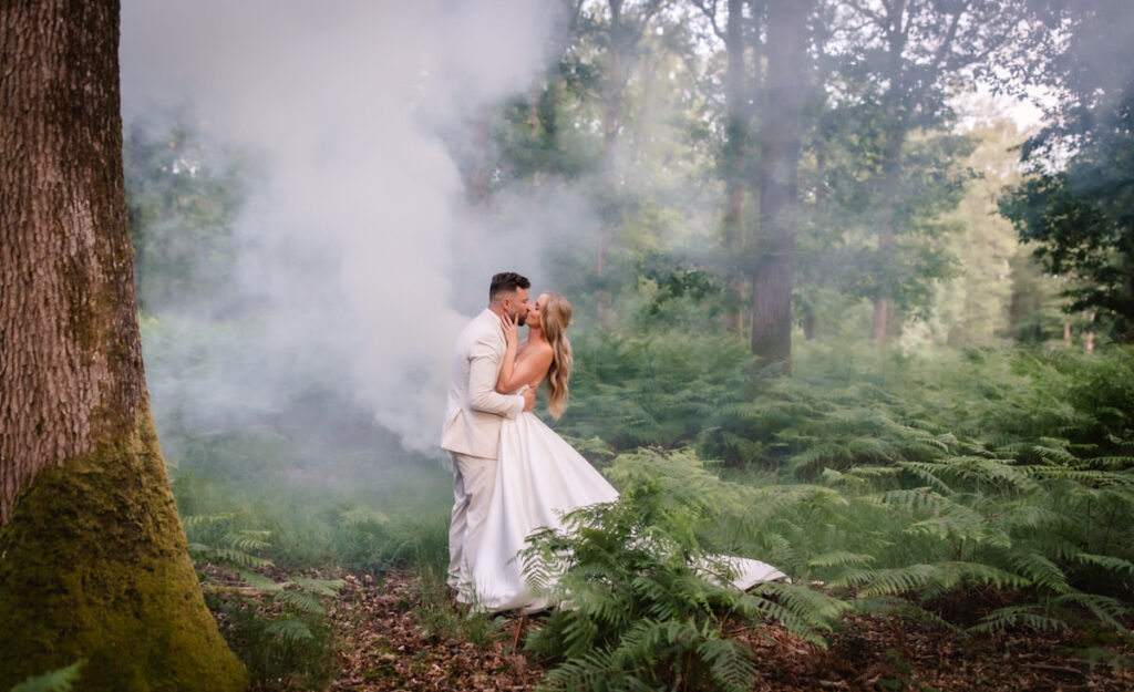 A bride and groom kiss in a lush green forest surrounded by ferns and mist, with sunlight filtering through the trees. The bride wears a white dress and the groom a light suit.