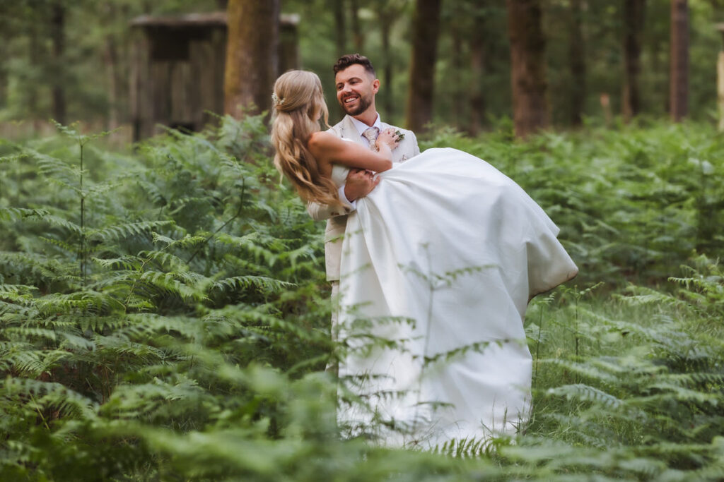 A groom in a light suit carries a smiling bride in a white wedding dress through lush green ferns in a forest, with trees and a rustic wooden structure in the background.