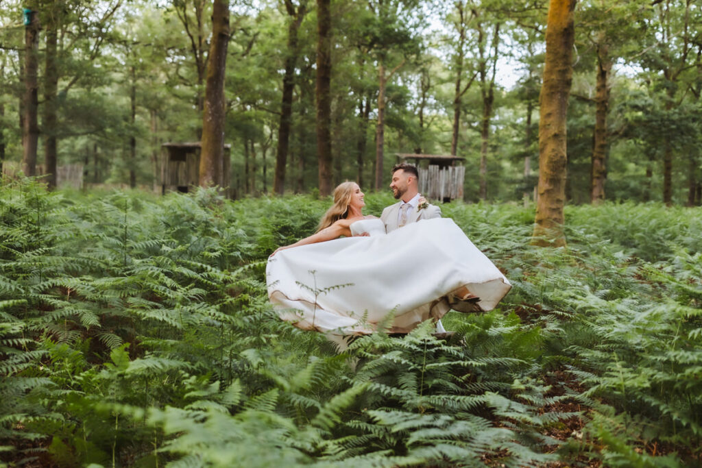 A bride and groom stand in a lush, green forest surrounded by ferns. The bride’s white dress billows outward as they gaze at each other, creating a romantic and dreamy atmosphere amid the tall trees.