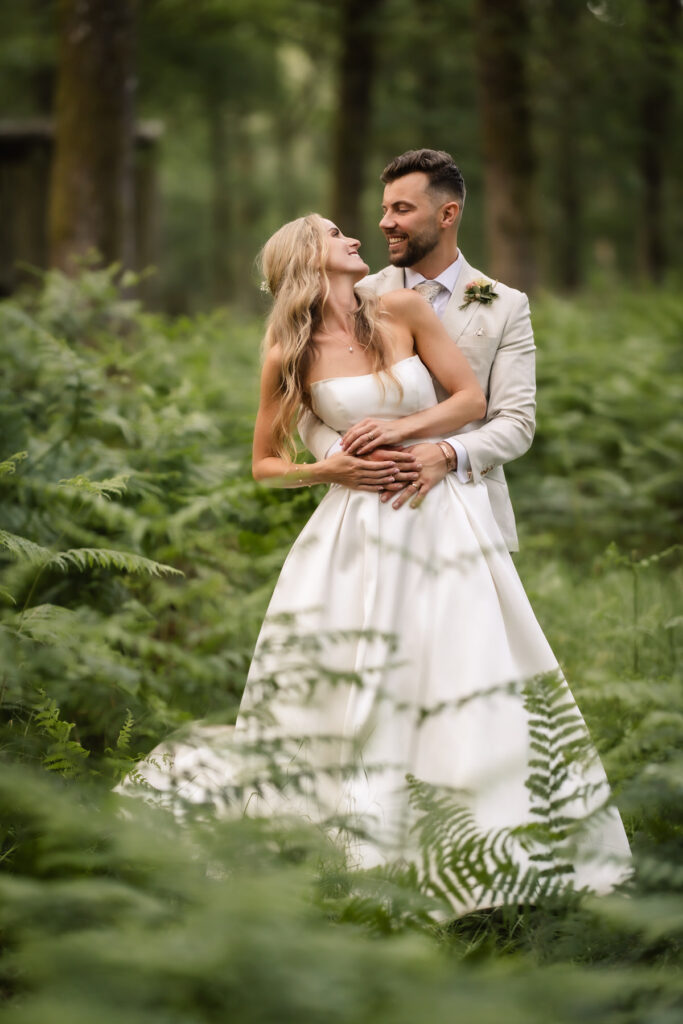 A bride in a white dress and a groom in a light suit stand embracing and smiling at each other in a lush, green forest filled with ferns.