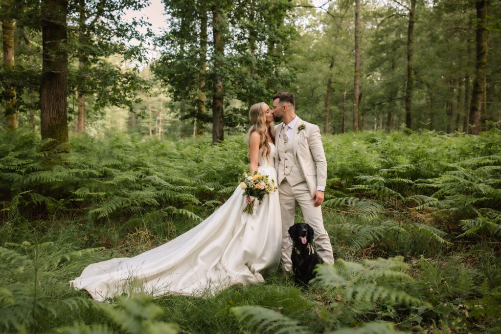 A bride and groom stand in a lush forest, sharing a kiss. The bride wears a long white gown and holds a bouquet, while a black dog sits at their feet, surrounded by green ferns and tall trees.