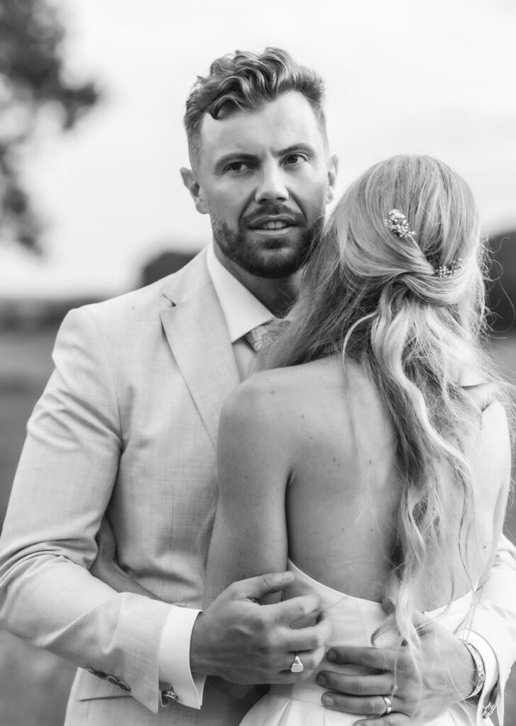 A man in a light suit embraces a woman in a backless dress with long wavy hair, standing outdoors. The photo is black and white, and the man looks toward the camera while the woman’s face is turned away.