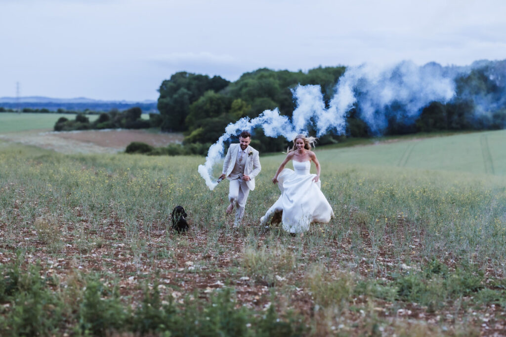 A bride and groom run joyfully through a field with a black dog, trailing blue smoke behind them. Lush green countryside and trees are visible in the background under a cloudy sky.