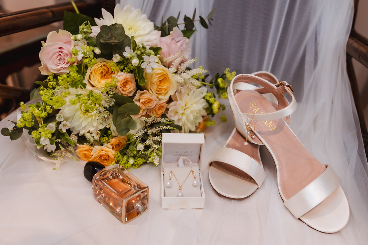 A bridal still life featuring a bouquet of pastel flowers, white sandals, a perfume bottle, and a jewelry box with pearl earrings, arranged on a white veil.