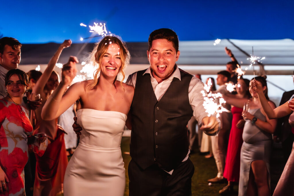 A joyful bride and groom walk arm in arm at night, holding sparklers, surrounded by cheering guests in colorful attire at an outdoor celebration.