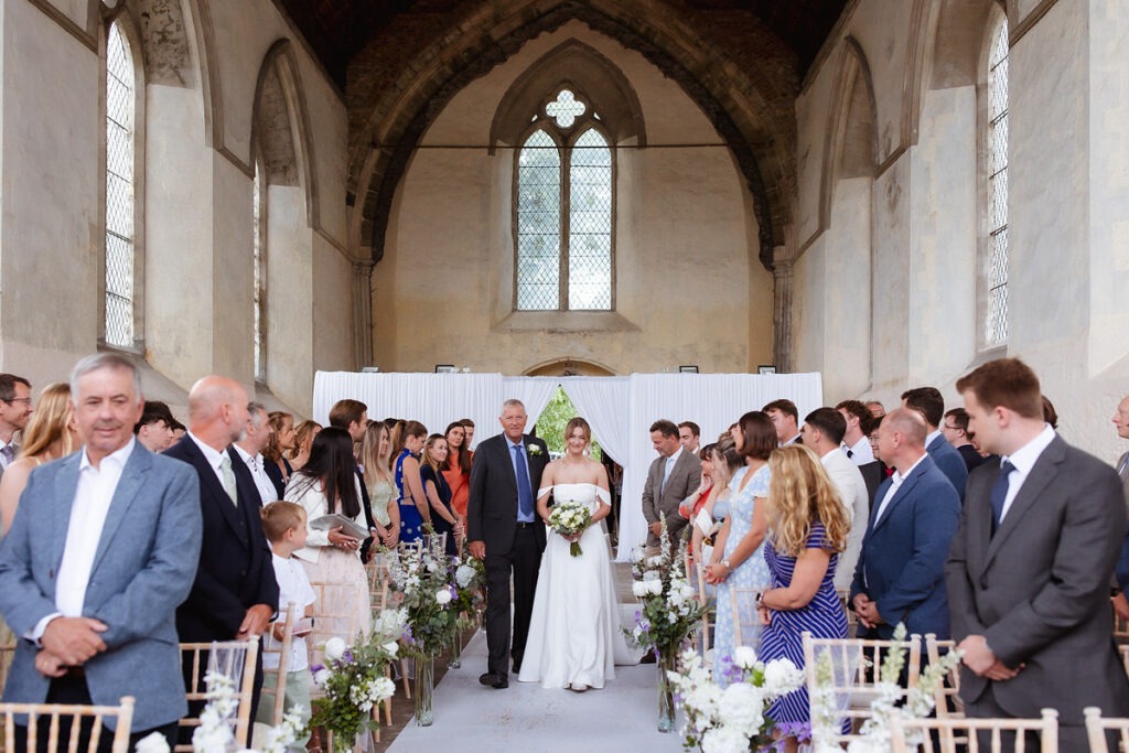 A bride in a white dress walks down the aisle with an older man in a suit inside a church, surrounded by seated and standing guests who are watching and smiling. The venue features tall windows and floral aisle decorations.