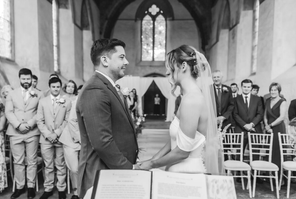 A bride and groom stand facing each other, holding hands at the altar in a chapel, surrounded by smiling guests. The scene is captured in black and white, with the officiant and a window in the background.