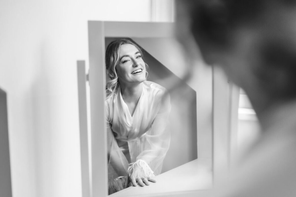 A woman in a white robe smiles while looking at her reflection in a mirror. The photo is black and white, and she appears joyful and relaxed. The focus is on her reflection, with her hand resting on a surface.