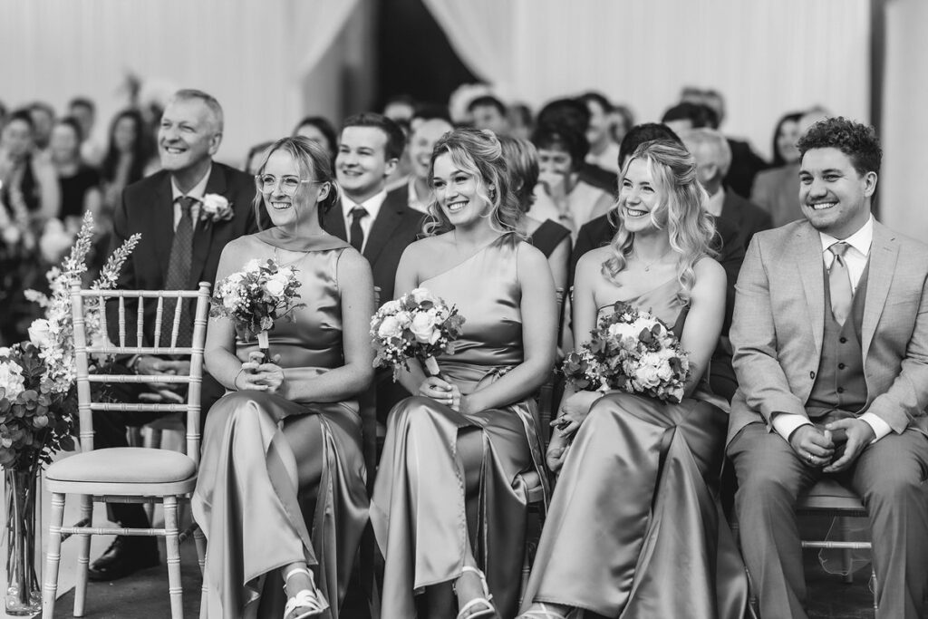 A group of bridesmaids in matching dresses and a groomsman in a suit sit smiling in the front row at a wedding ceremony, with guests seated behind them.