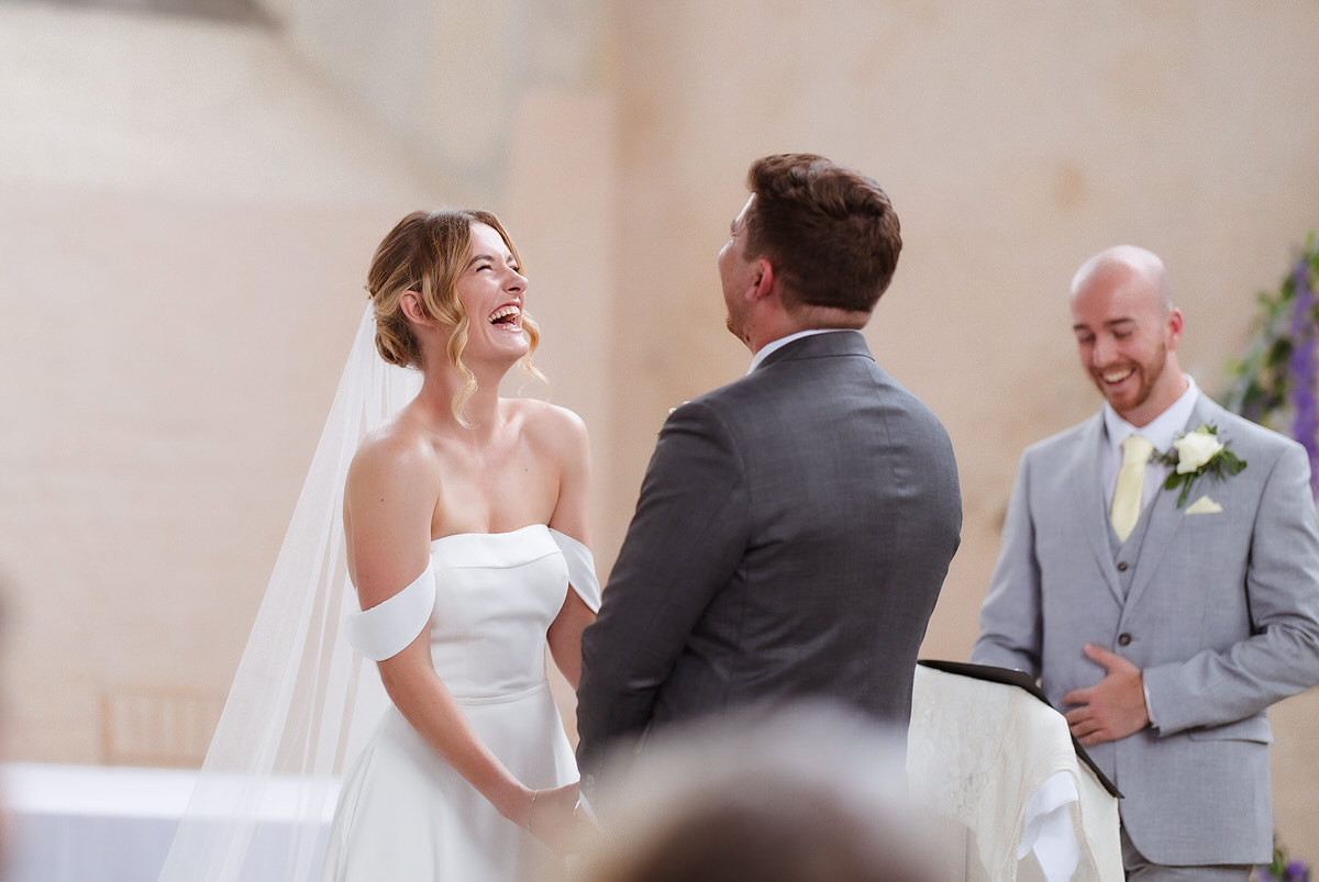 A bride in an off-shoulder white gown and a groom in a gray suit laugh joyfully during their wedding ceremony, with a smiling officiant standing beside them.