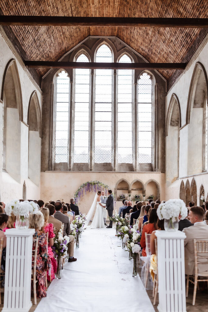 A bride and groom stand at the altar in a grand, high-ceilinged hall with tall arched windows, surrounded by seated guests and floral decorations along the aisle.
