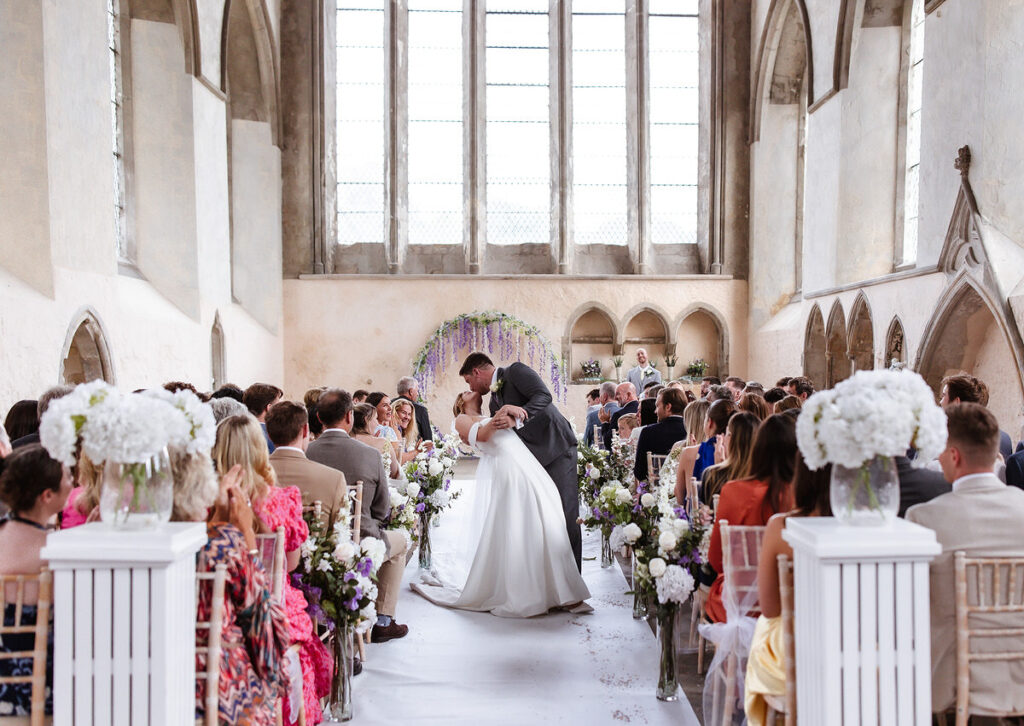 A bride and groom share a kiss at the altar during their wedding ceremony in a bright, flower-filled church. Guests sit in rows, watching the couple under tall arched windows that fill the space with natural light.