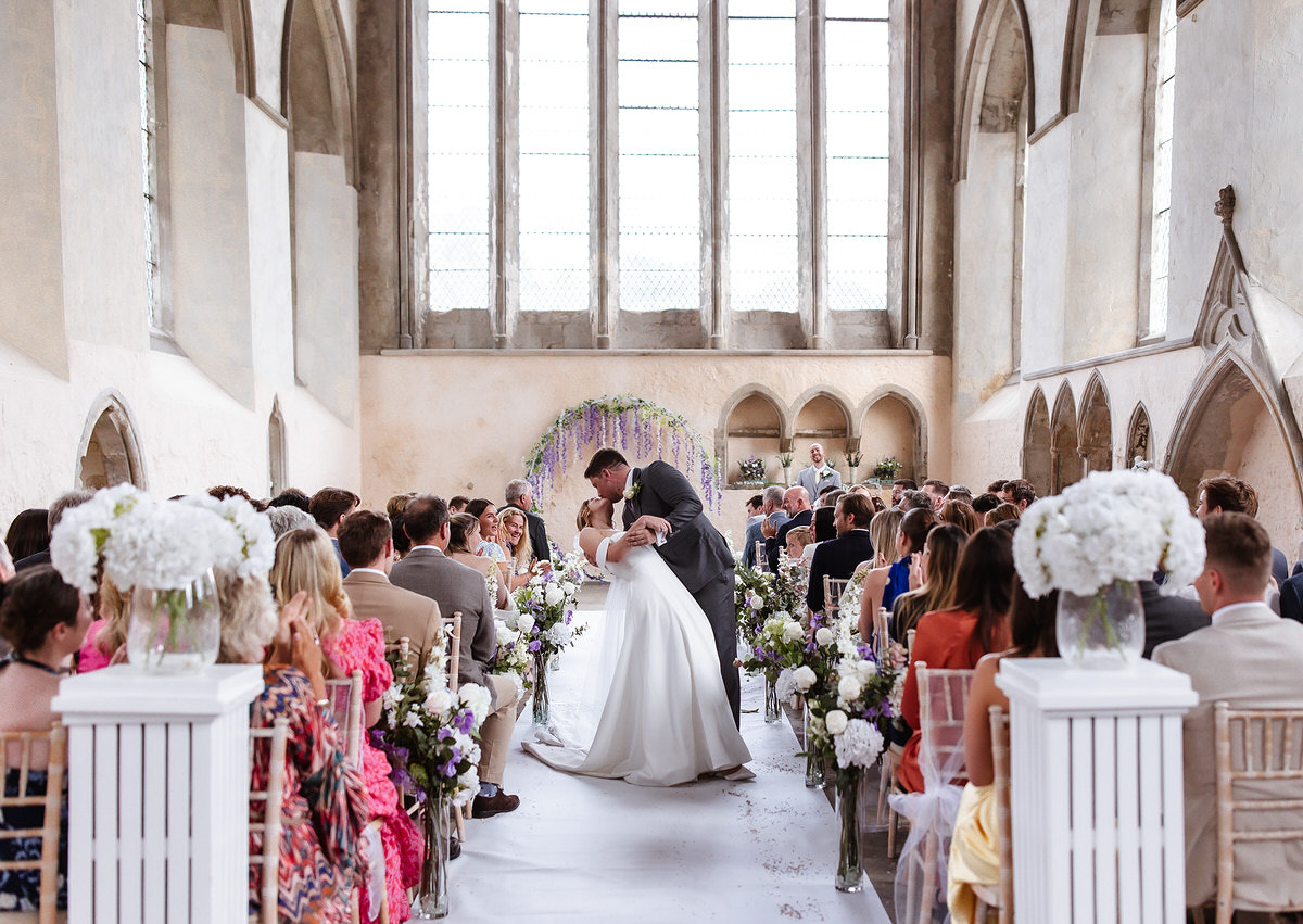 A bride and groom share a kiss at the end of the aisle in a bright, flower-filled church, surrounded by seated guests watching their wedding ceremony.