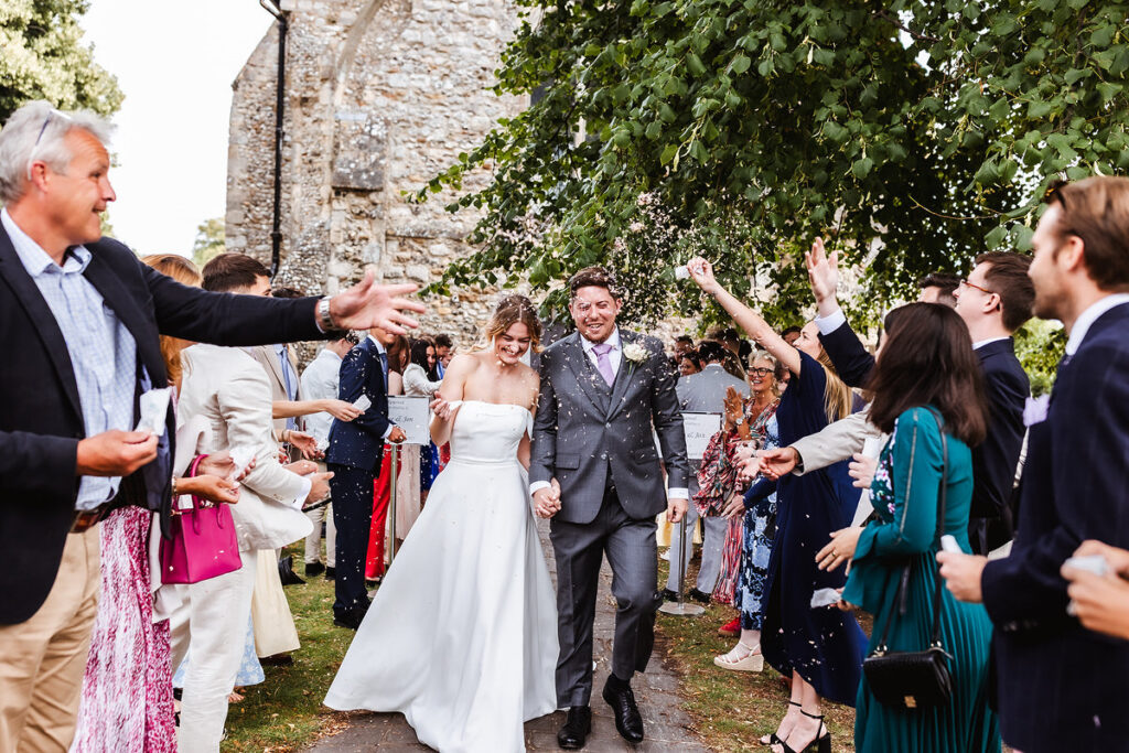 A bride and groom walk hand in hand outside as wedding guests cheer and throw confetti. Both are smiling, surrounded by friends and family in festive attire near a stone building and leafy trees.
