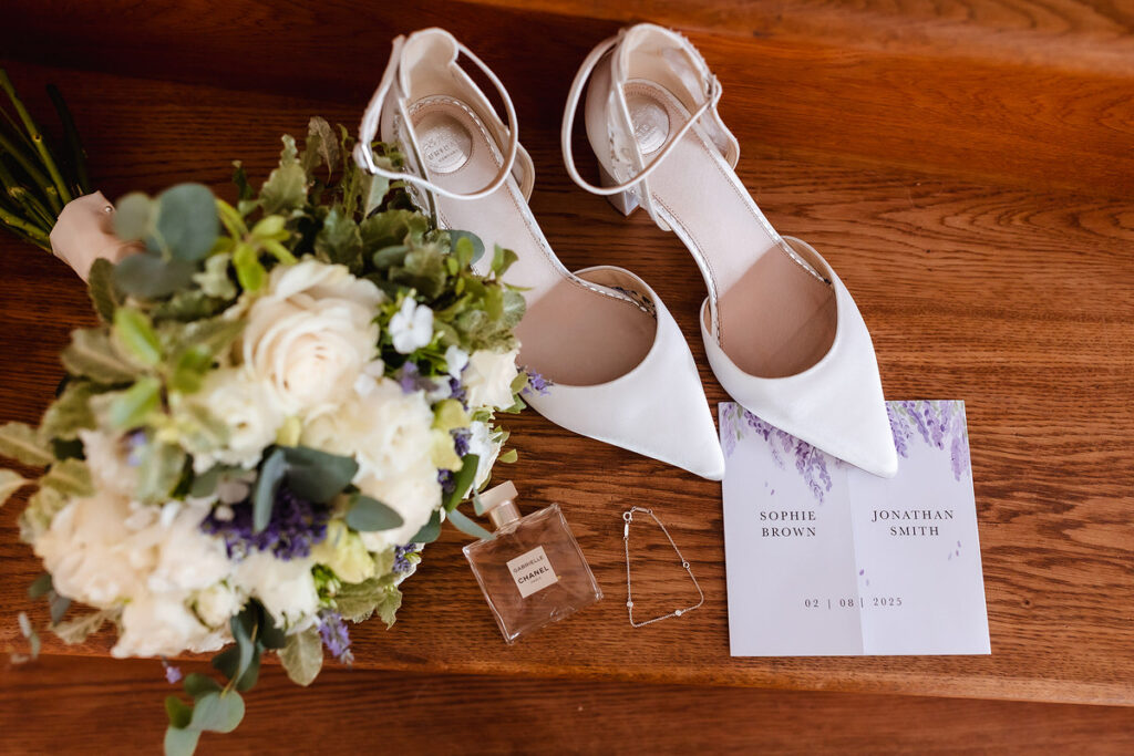 White bridal shoes, a bouquet of white and green flowers, a perfume bottle, jewelry, and a wedding invitation with the names Sophie Brown and Jonathan Smith are arranged on wooden stairs.
