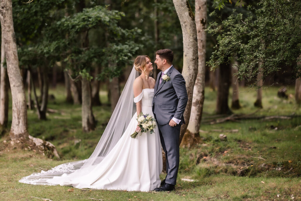 A bride in a white dress and veil stands smiling with her groom in a gray suit, holding a bouquet, surrounded by trees in a forest setting.