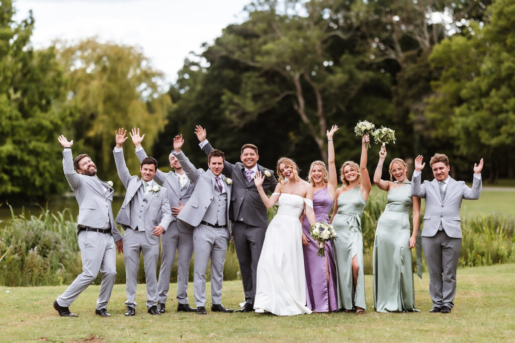 A wedding party of five men in grey suits and four women in pastel dresses stand outdoors on grass, smiling and cheering with their arms raised, surrounded by greenery and trees in the background.
