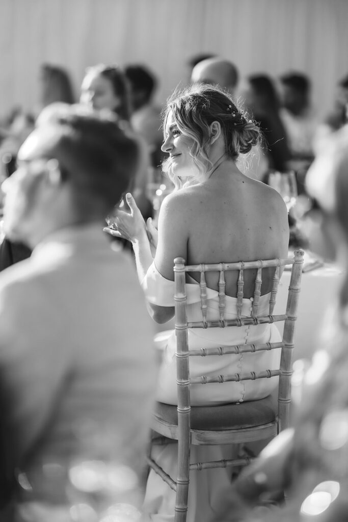 A woman in a strapless dress sits on a wooden chair at an event, turning to smile. Other people are seated around her, and the atmosphere is bright and lively. The photo is in black and white.